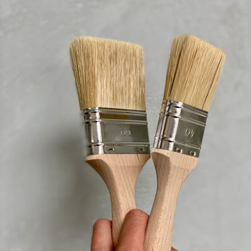 Zwei Pinsel mit Holzgriffen vor einer grauen Wand, die das klassische Werkzeug eines Malers sind.