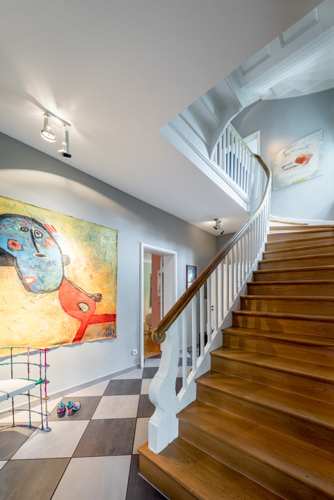Großzügiges Treppenhaus mit geschwungener Holztreppe, weißem Geländer und wohngesunden, glatten Wänden in Mineralfarbe – kombiniert mit Kunst und stilvoller Beleuchtung für ein gesundes und elegantes Wohnambiente.
