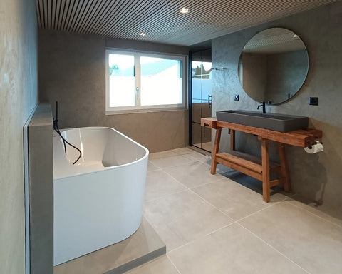 Modernes Badezimmer mit fugenlos gestalteten Wänden aus Beton Ciré, freistehender Badewanne, großem Rundspiegel und Holzwaschtisch.