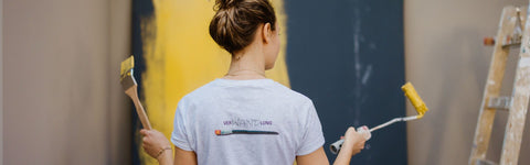 Eine Malermeisterin in grauem T-Shirt die einen Pinsel und eine Rolle in den Händen hält vor einer gelb und anthrazit gestrichenen Wand.